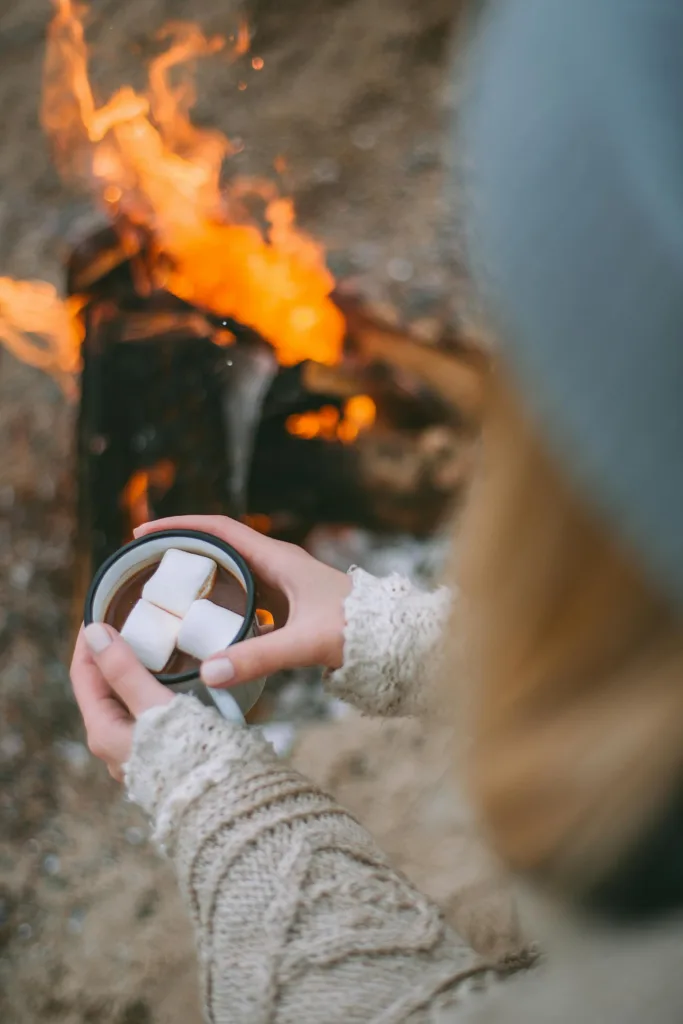 Cozy winter scene with a woman holding hot chocolate near a campfire, perfect for lifestyle imagery.