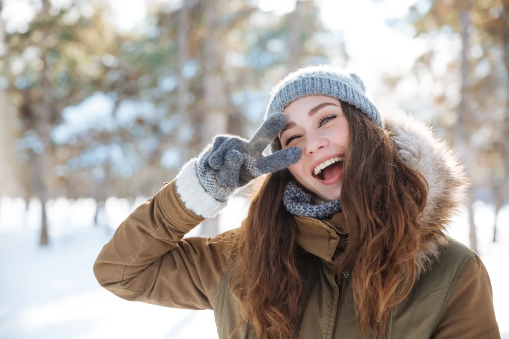 Woman in the snow wearing a warm jacket, beanie and gloves making a peace sign.