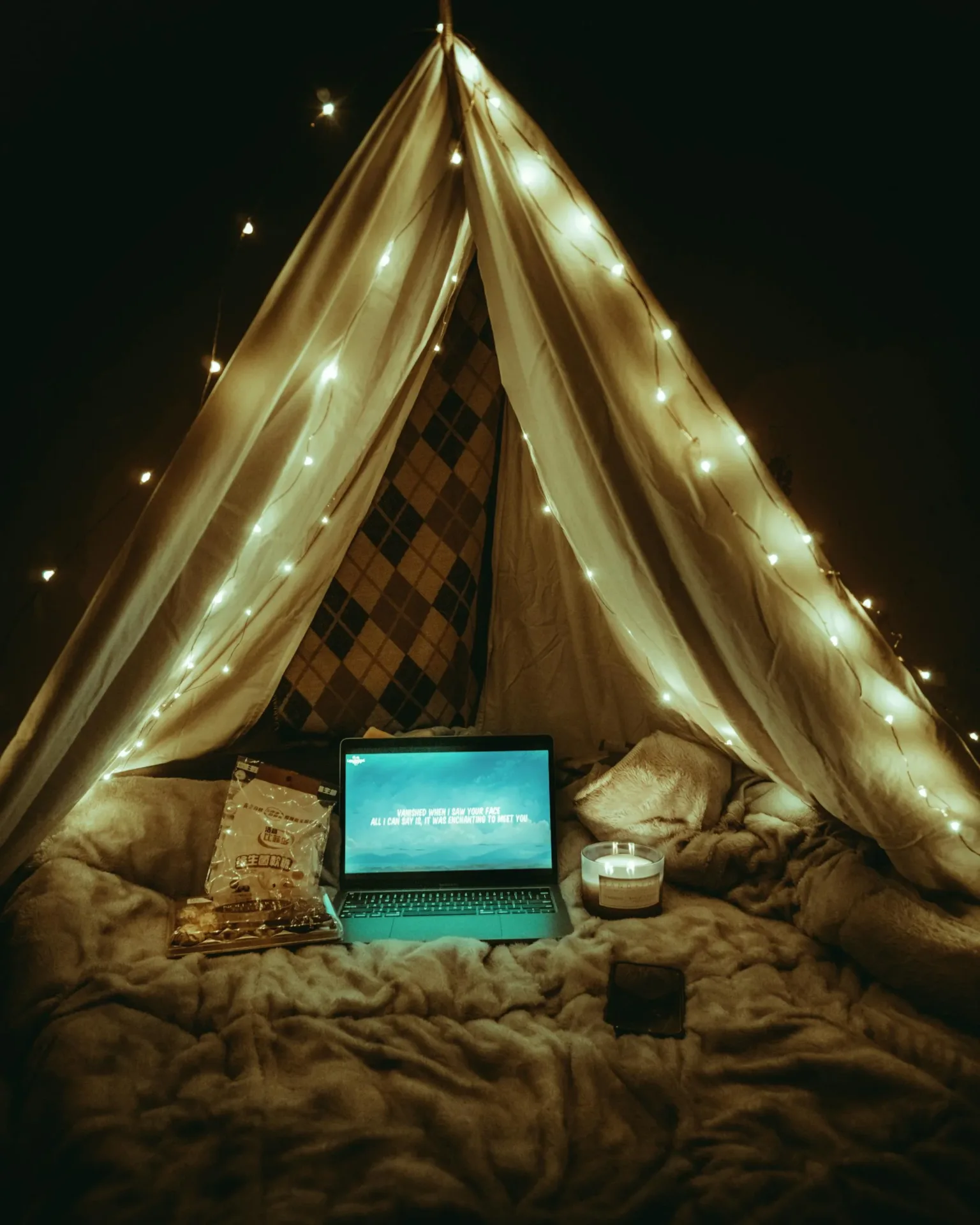 A relaxing indoor tent setup with fairy lights, laptop, and snacks for a cozy movie night.