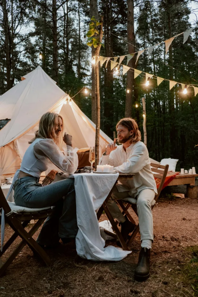 A couple enjoys a romantic dinner outside a tent in the woods, under string lights.