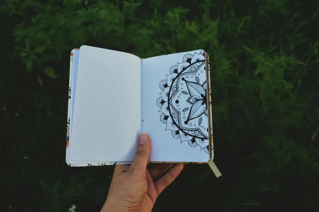 A hand holding an open journal with a detailed mandala drawing outdoors.