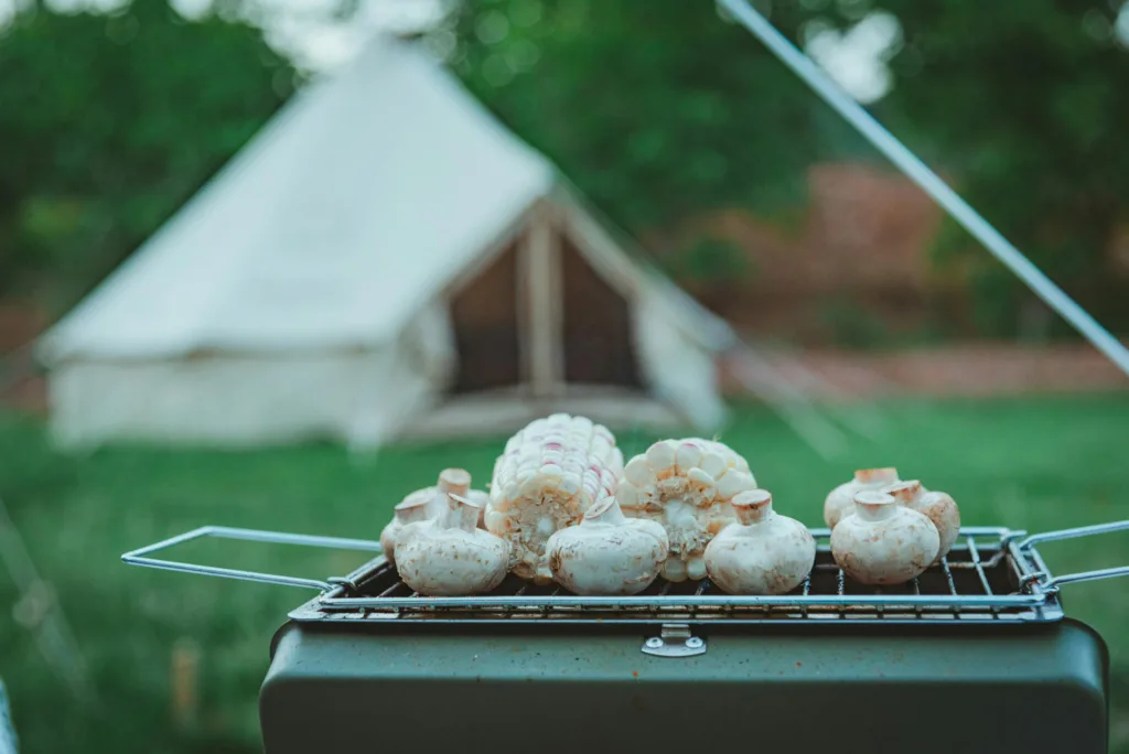 Outdoor grilling with mushrooms and corn near a tent, perfect for camping enthusiasts.