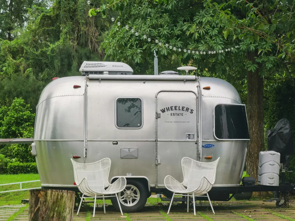 A vintage Airstream caravan parked in a lush Singapore garden with two modern white chairs.