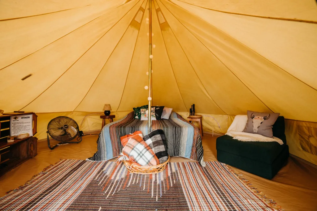 Bed with colorful blanket near comfortable armchair with cushions placed in yellow spacious tent with colorful carpet and decorated pole