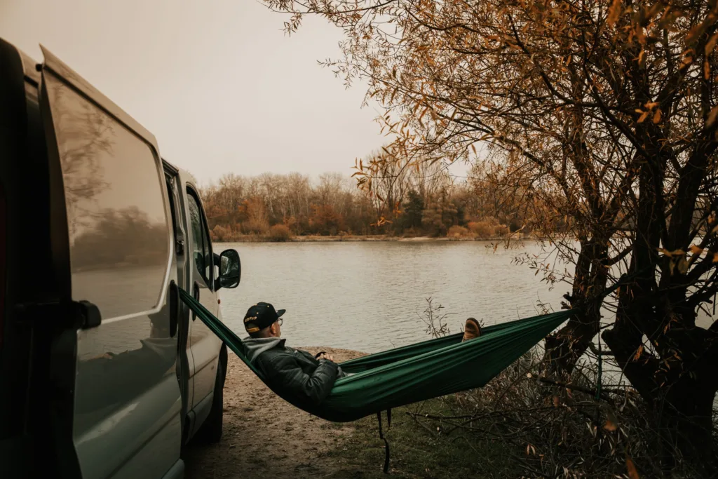 A tranquil autumn scene with a person relaxing in a hammock next to a van by the river.