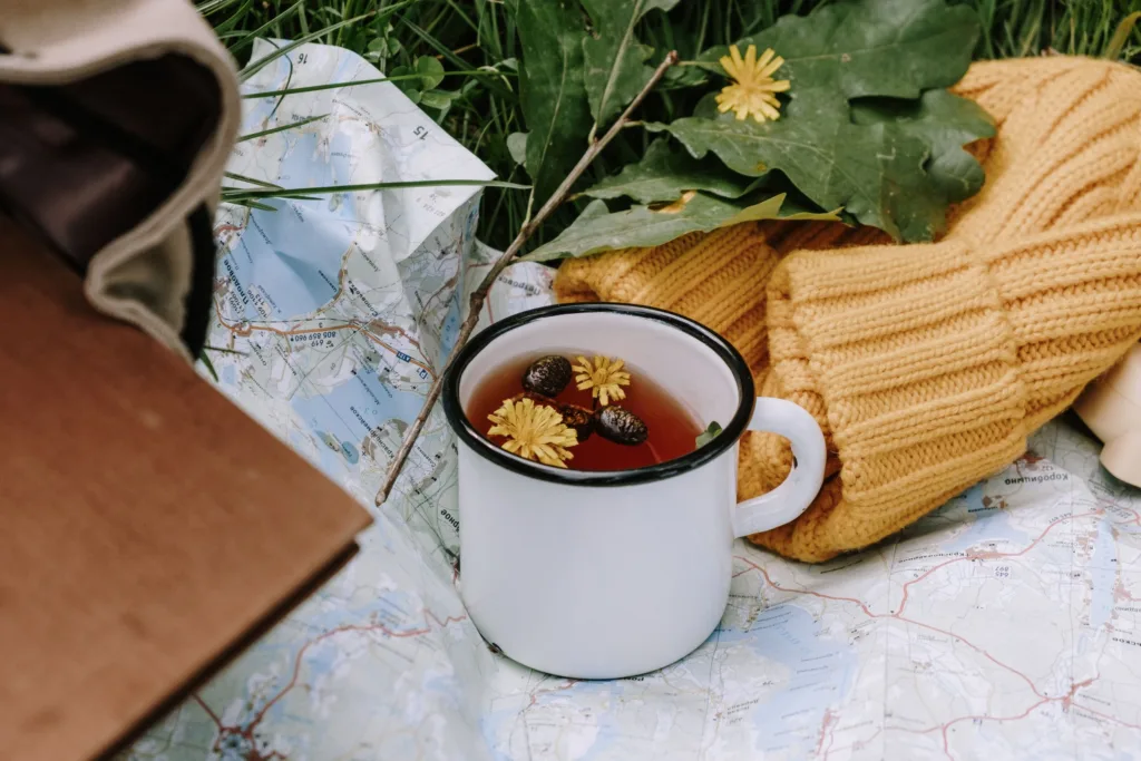 A white ceramic mug of tea with yellow flowers, on a map outdoors alongside a yellow knitted beanie.