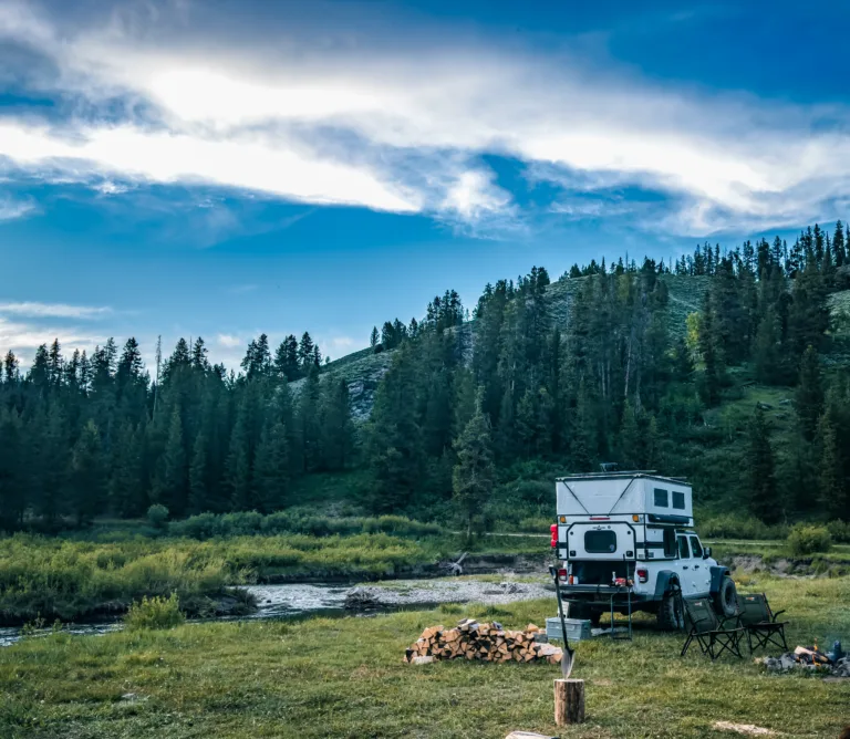 Jeep Gladiator with a truck camper parked at a dispersed camping spot with a river in the background.