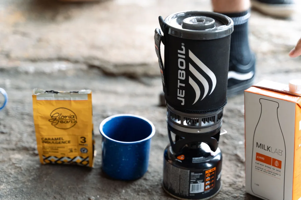 A close-up of a camp stove alongside coffee and milk preparation setup outdoors.