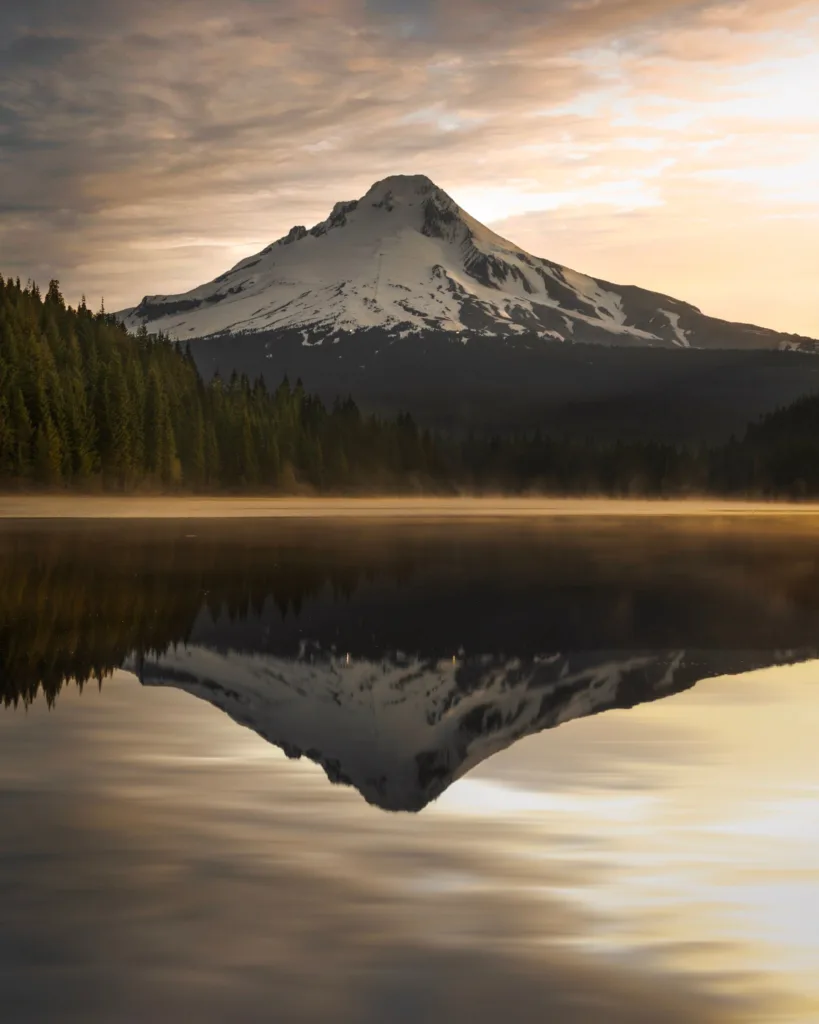 Idyllic sunrise view of snow-capped mountain reflected in a tranquil lake, surrounded by lush pine trees.